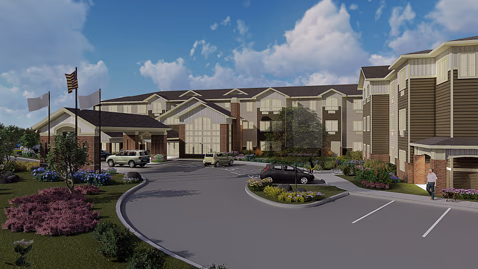 Exterior view of a large senior living facility building with a covered entrance, several parked cars, landscaped greenery, and three flagpoles with flags including an American flag under a partly cloudy sky.