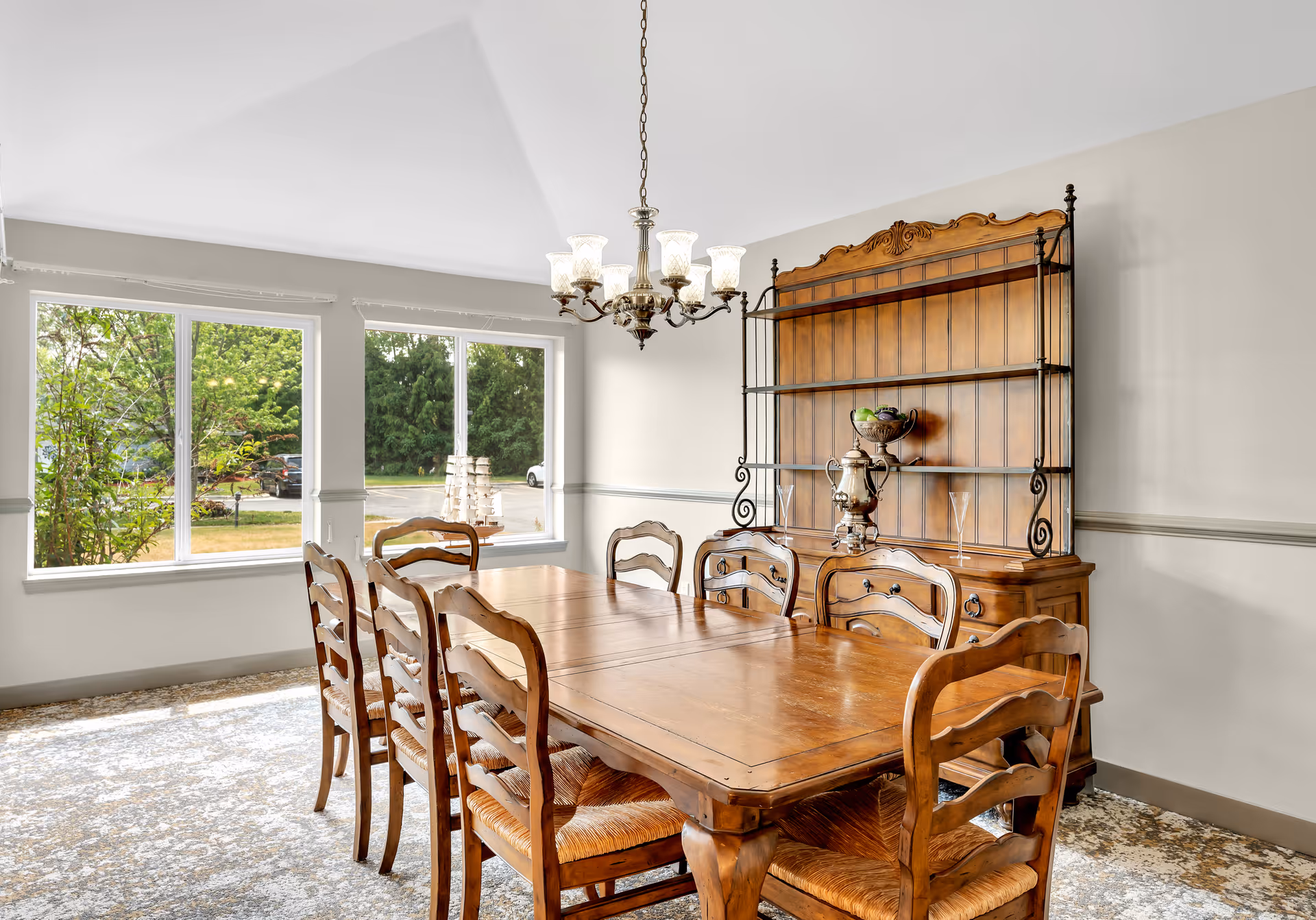 A dining room with a large wooden table surrounded by eight matching wooden chairs with woven seats. A wooden hutch with metal accents stands against the wall, displaying a decorative bowl with fruit and a silver tea set. A chandelier with multiple glass shades hangs above the table. Large windows let in natural light and show greenery and parked cars outside.