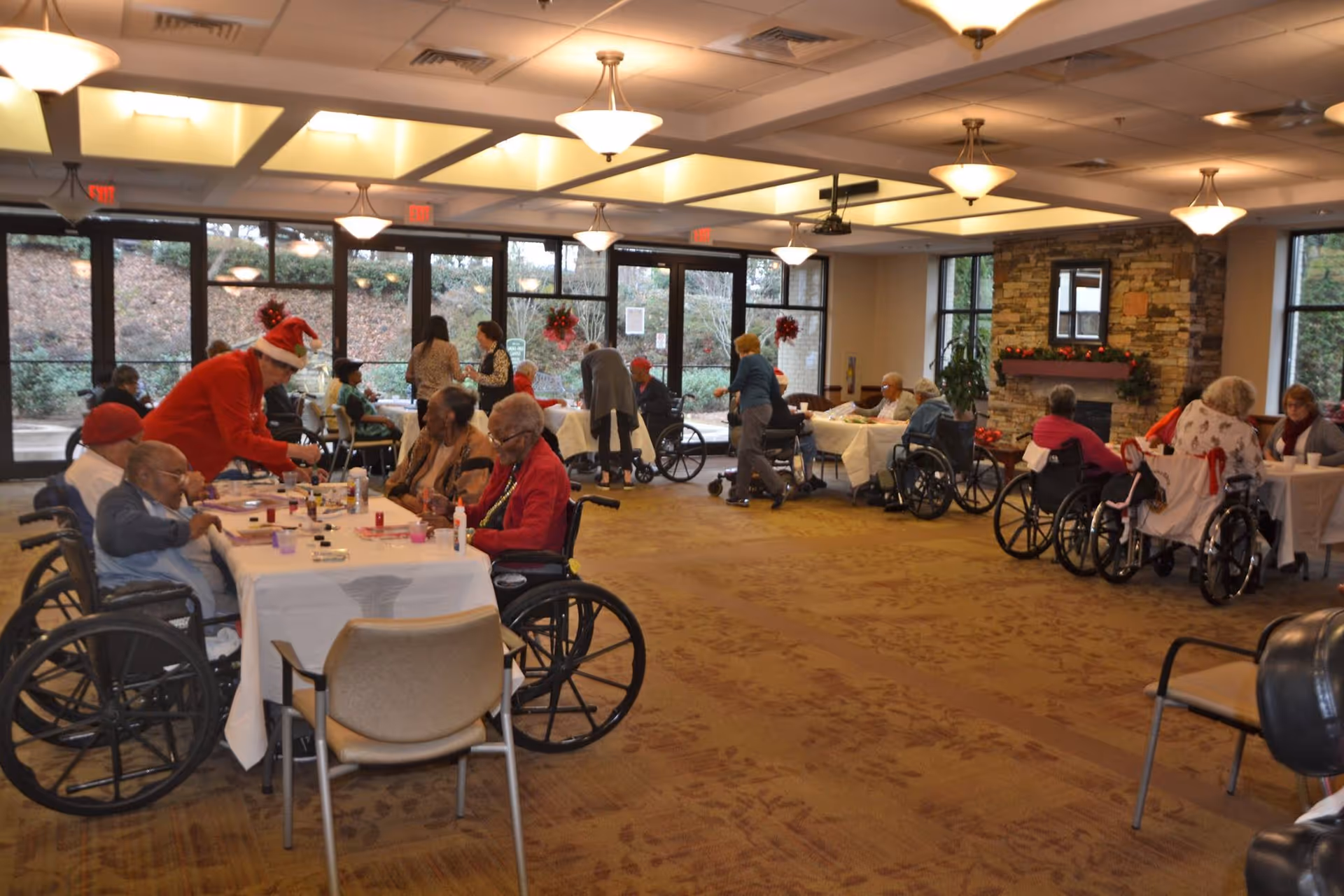 A group of elderly individuals, many in wheelchairs, seated around tables in a spacious, well-lit common room with large windows. Staff members are assisting and interacting with the residents, some wearing festive holiday attire including Santa hats. The room has a warm, inviting atmosphere with beige carpet and ceiling lights.