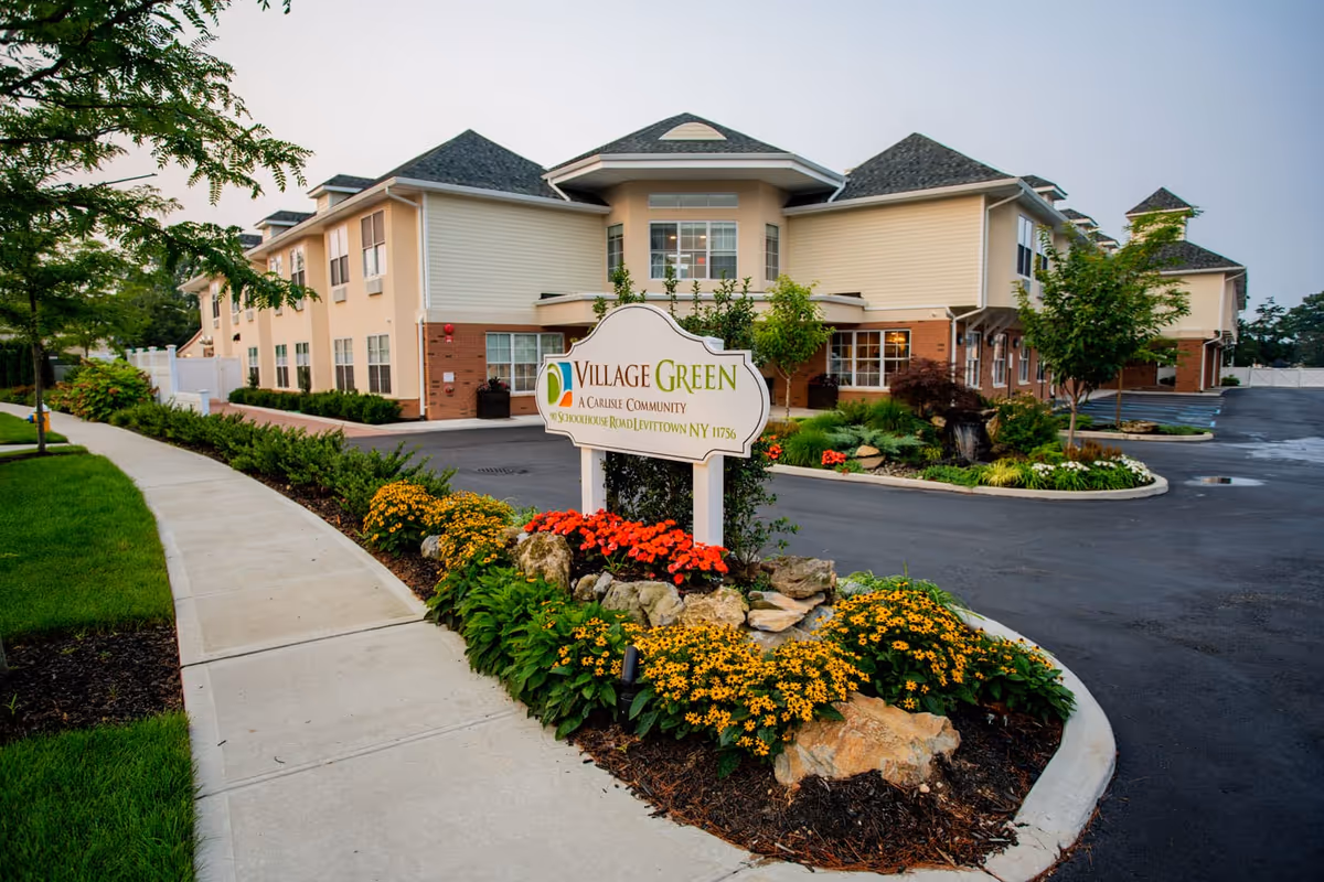 Front exterior of the Village Green senior living building with a landscaped entrance sign and flowerbeds.