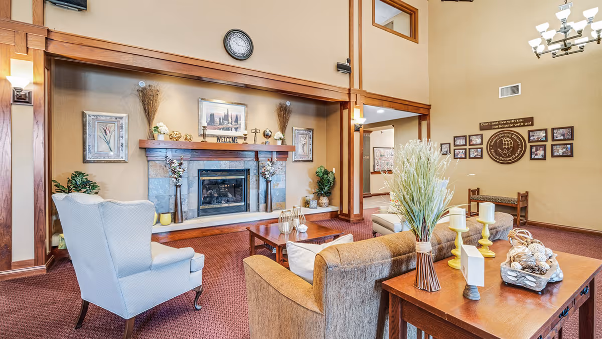 A cozy senior living facility common area with a fireplace surrounded by decorative vases and framed artwork. The room features comfortable seating including a beige armchair and a brown sofa, a wooden coffee table, and a side table with candles and a decorative basket. The walls are painted beige with wooden trim, and there is a clock above the fireplace. On the far wall, there are framed photos and a sign that reads 'Don't just live with us - participate with us!'