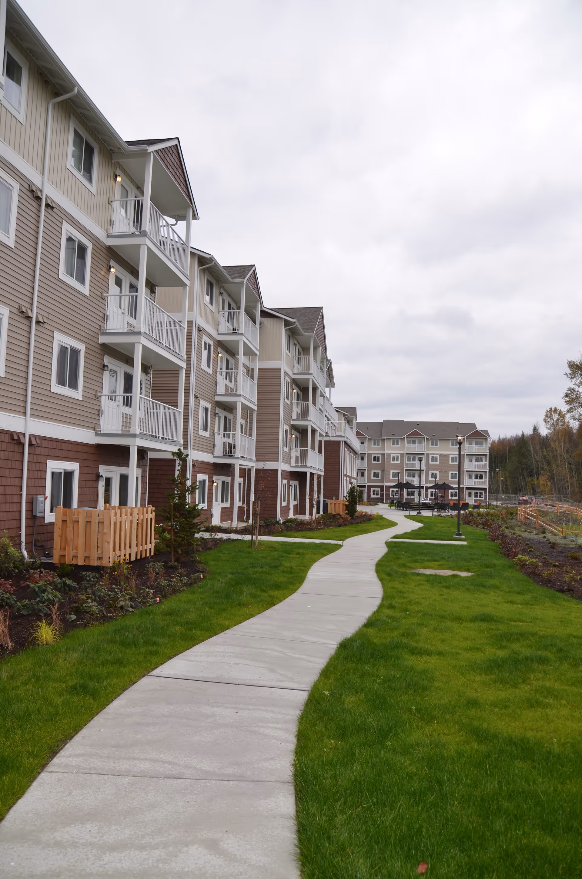 Multi-story residential building with balconies next to a curved sidewalk and green lawns under an overcast sky.