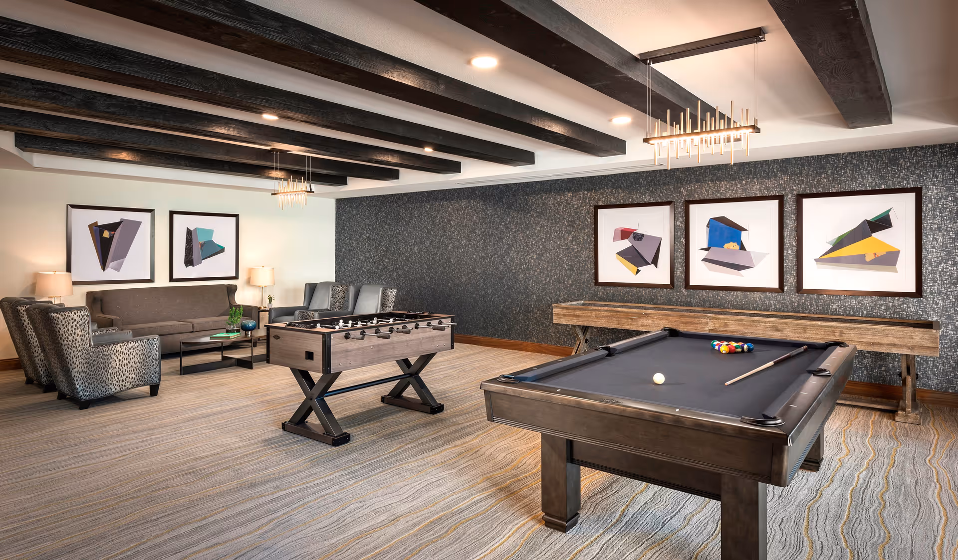 A communal game room with a pool table, foosball table, seating area, and framed artwork beneath exposed ceiling beams.