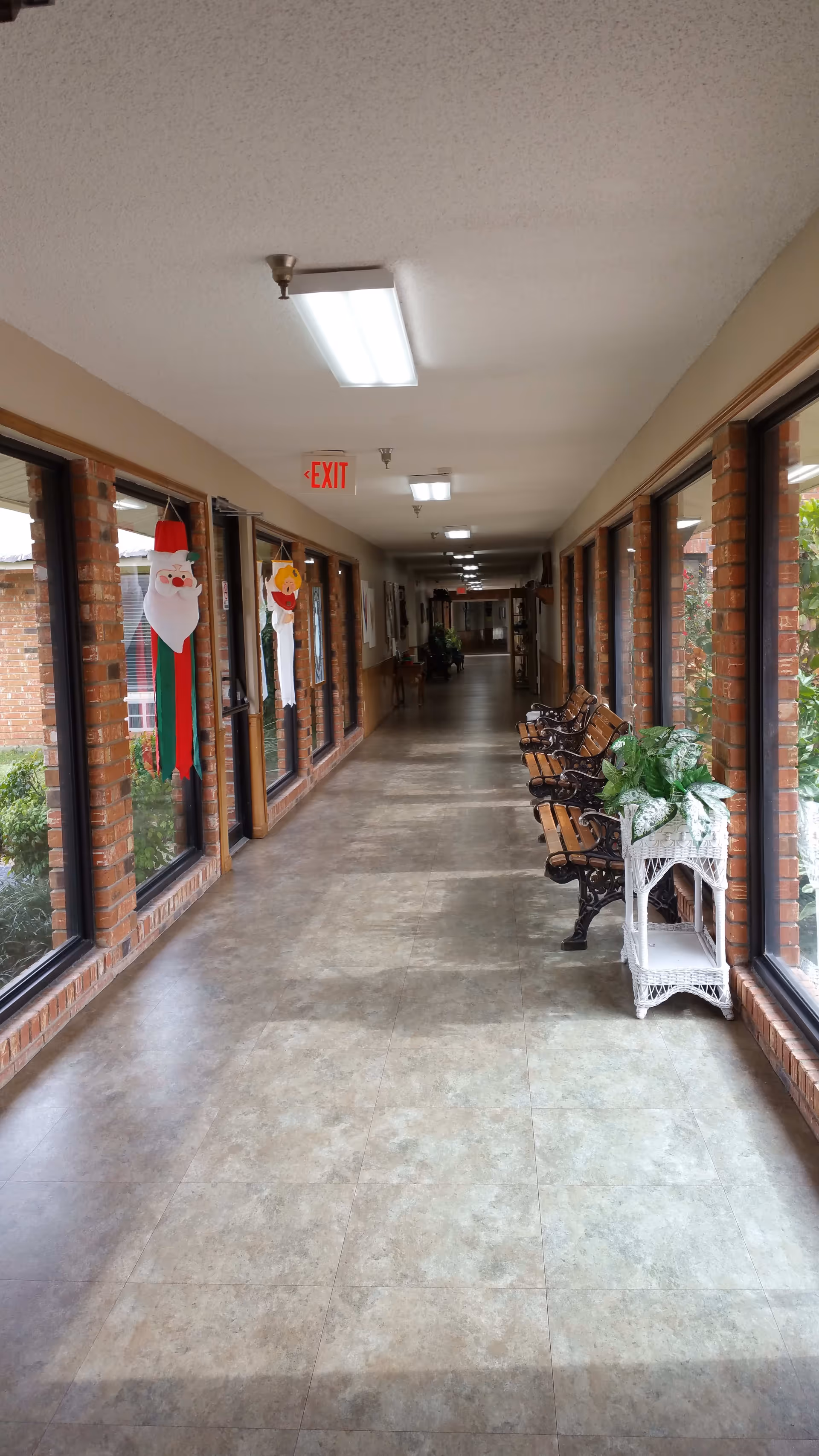 A long indoor hallway with large windows on the right side showing outdoor greenery. The left side has brick walls with windows and holiday decorations including a Santa Claus and a snowman hanging. There are benches and a white wicker plant stand with green plants along the right side. The ceiling has fluorescent lights and an exit sign is visible in the distance.