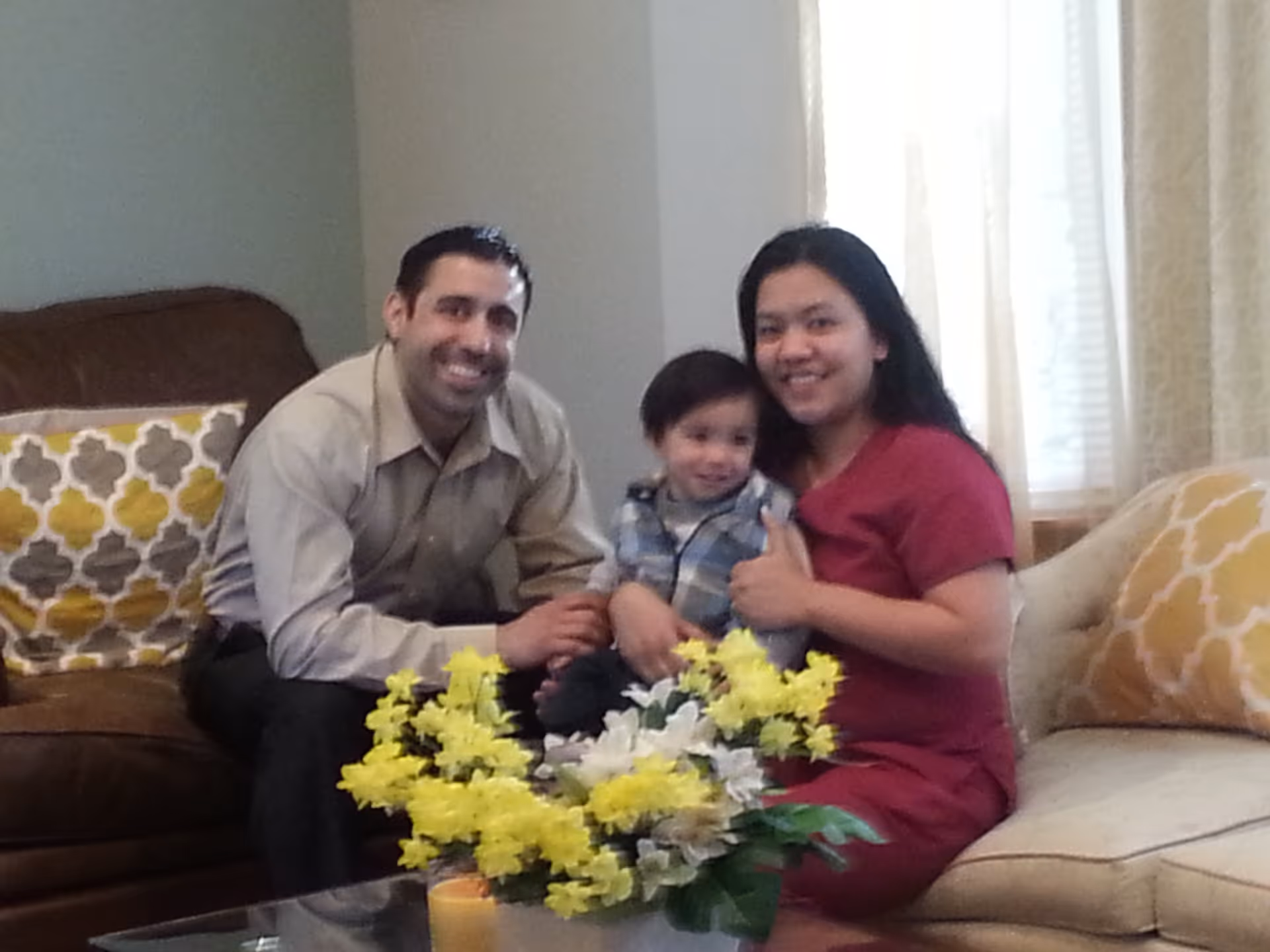 A family of three sitting together on sofas in a living room. The man is on the left on a brown sofa, the woman is on the right on a beige sofa holding a young child on her lap. There is a glass coffee table in front of them with a bouquet of yellow and white flowers. The room has light-colored curtains and decorative pillows on the sofas.