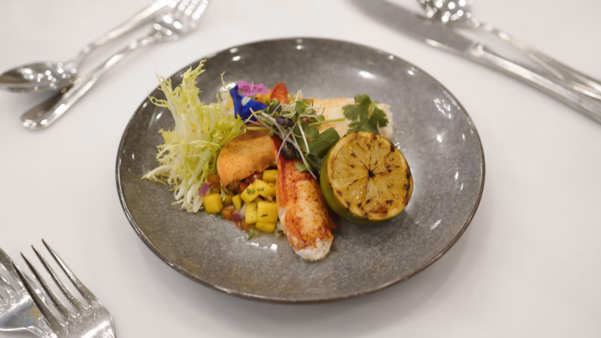 A gourmet dish on a gray plate featuring a grilled lime half, a piece of cooked seafood, a small portion of diced mango salsa, a leafy green garnish, and microgreens, set on a white tablecloth with silver cutlery around the plate.