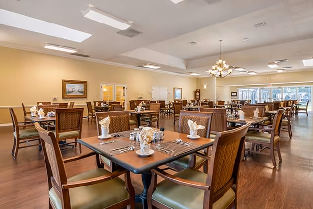 A spacious dining room in a senior living facility with multiple wooden tables and chairs arranged neatly. Each table is set with white napkins folded elegantly, cups, and silverware. The room has a warm beige color on the walls, a chandelier hanging from the ceiling, and large windows allowing natural light to fill the space.