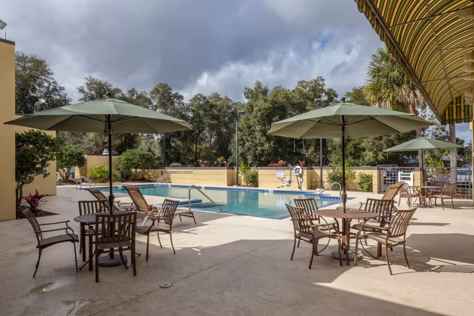 Outdoor pool area with several round tables and chairs under large green umbrellas. The pool is surrounded by a concrete deck and some greenery, with trees in the background under a partly cloudy sky.