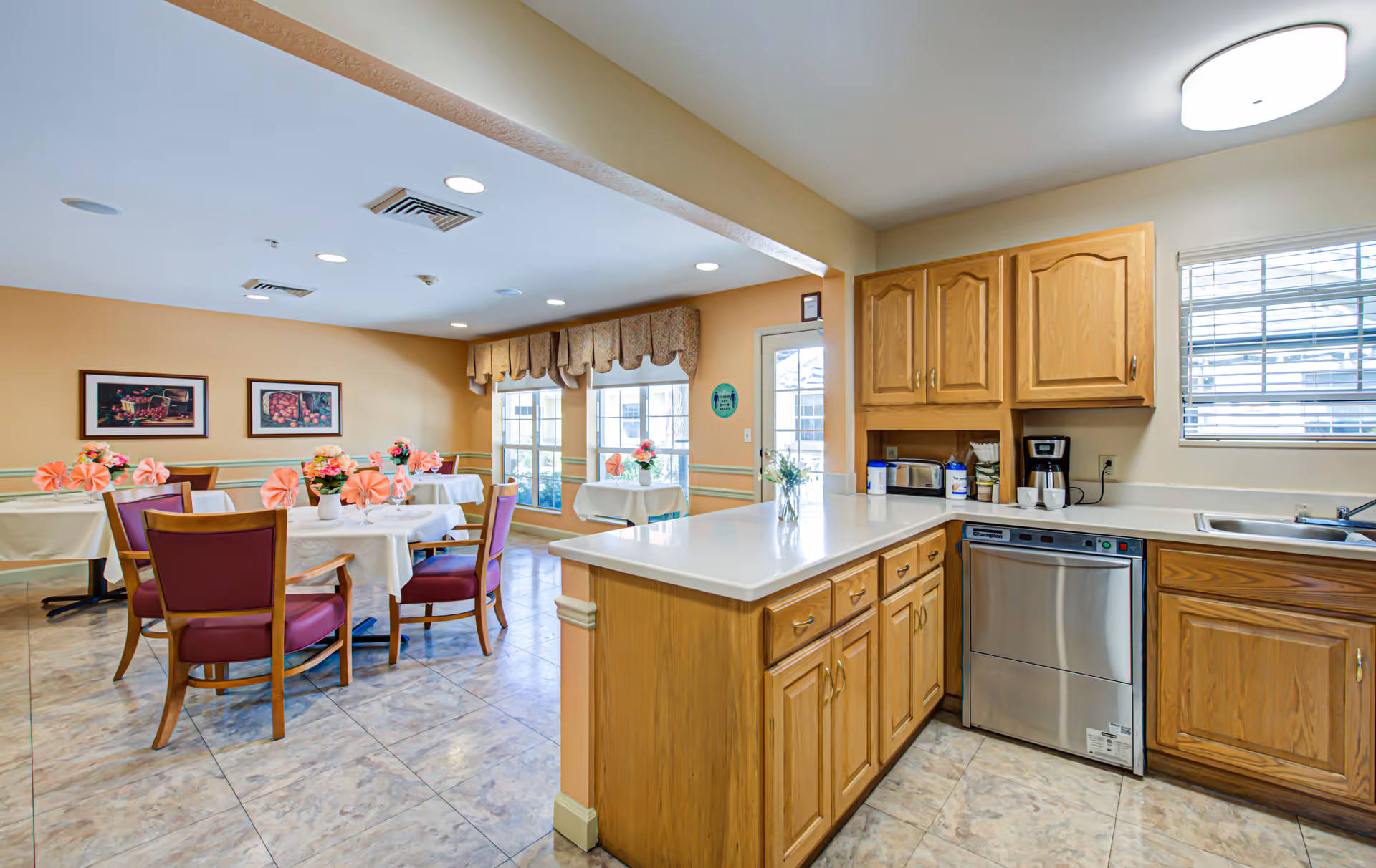 A bright dining area and kitchen in a senior living facility. The dining area has round tables covered with white tablecloths, each decorated with pink napkins and flower centerpieces. The kitchen features wooden cabinets, a dishwasher, a coffee maker, a toaster, and a sink under a window with blinds. The room has tiled floors and large windows letting in natural light.