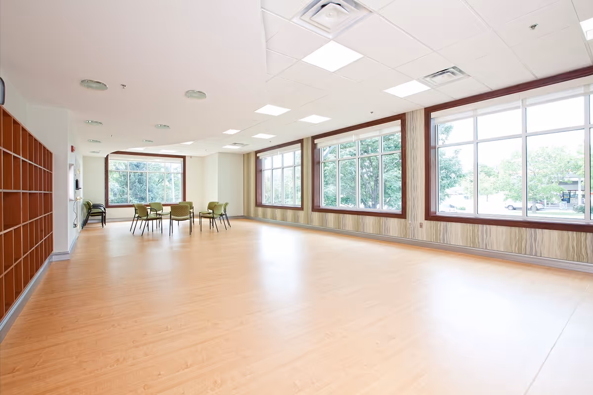 A spacious, well-lit room with large windows along two walls allowing natural light to fill the space. The room has light wood flooring, a white ceiling with recessed lighting, and a set of green chairs arranged around a table near the windows. There are wooden cubby shelves along one wall.