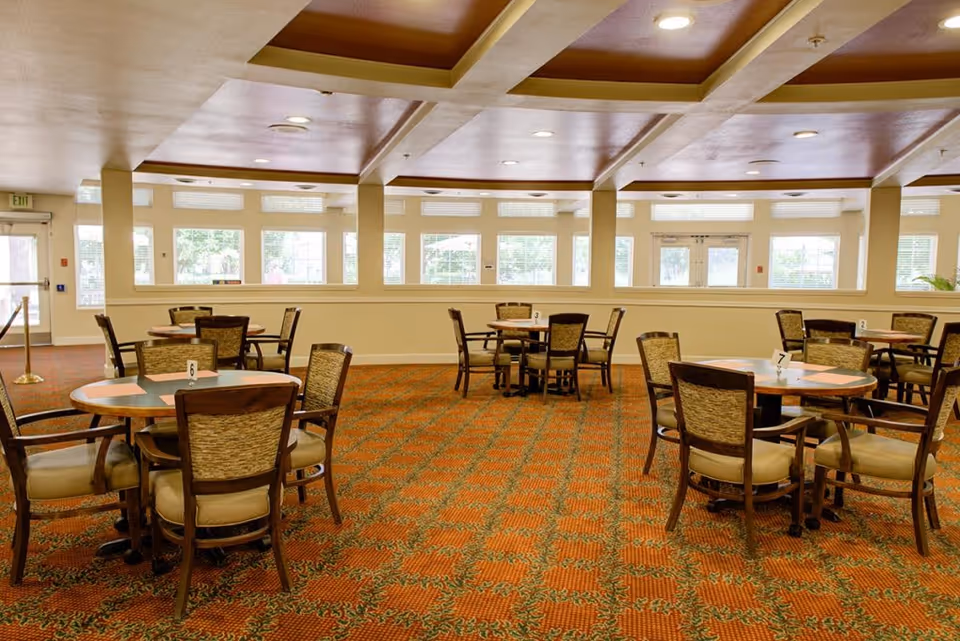A spacious dining room with multiple round tables and cushioned chairs arranged neatly on a patterned carpet. Large windows along the walls allow natural light to fill the room, and the ceiling features recessed lighting and a coffered design.