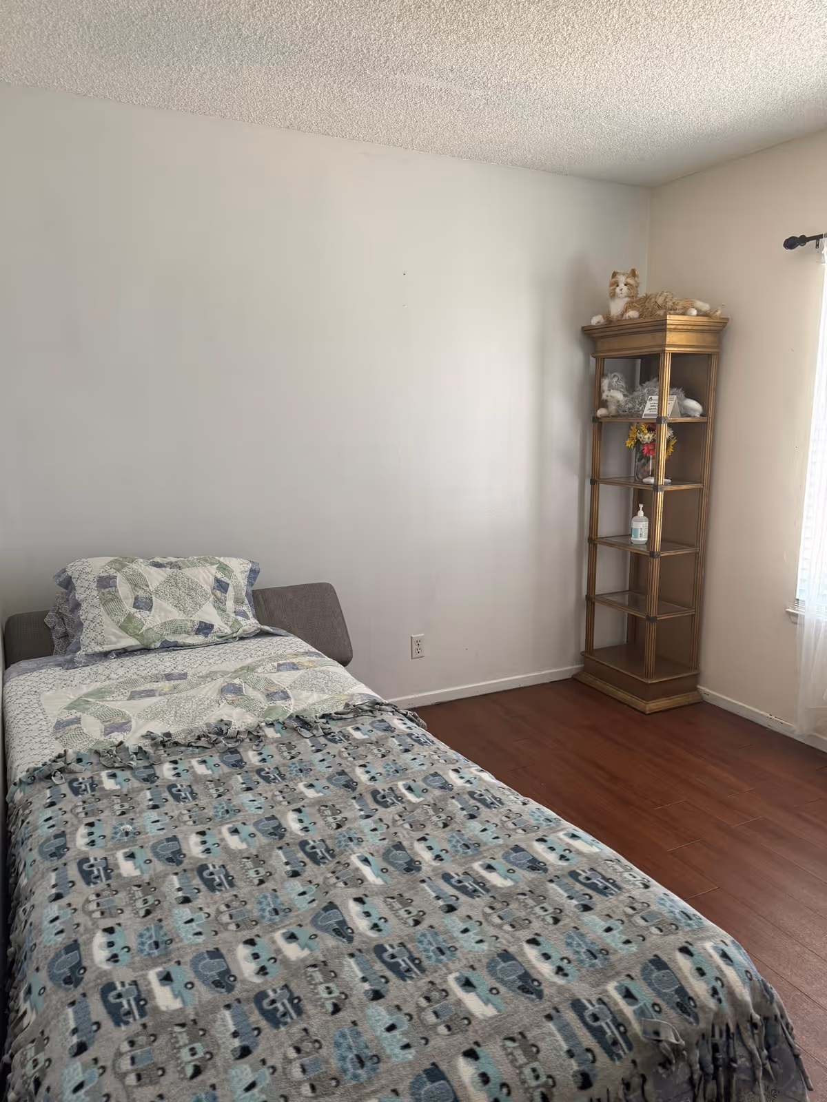 A small bedroom with a single bed covered by patterned quilts and a tall glass display shelf beside a window.