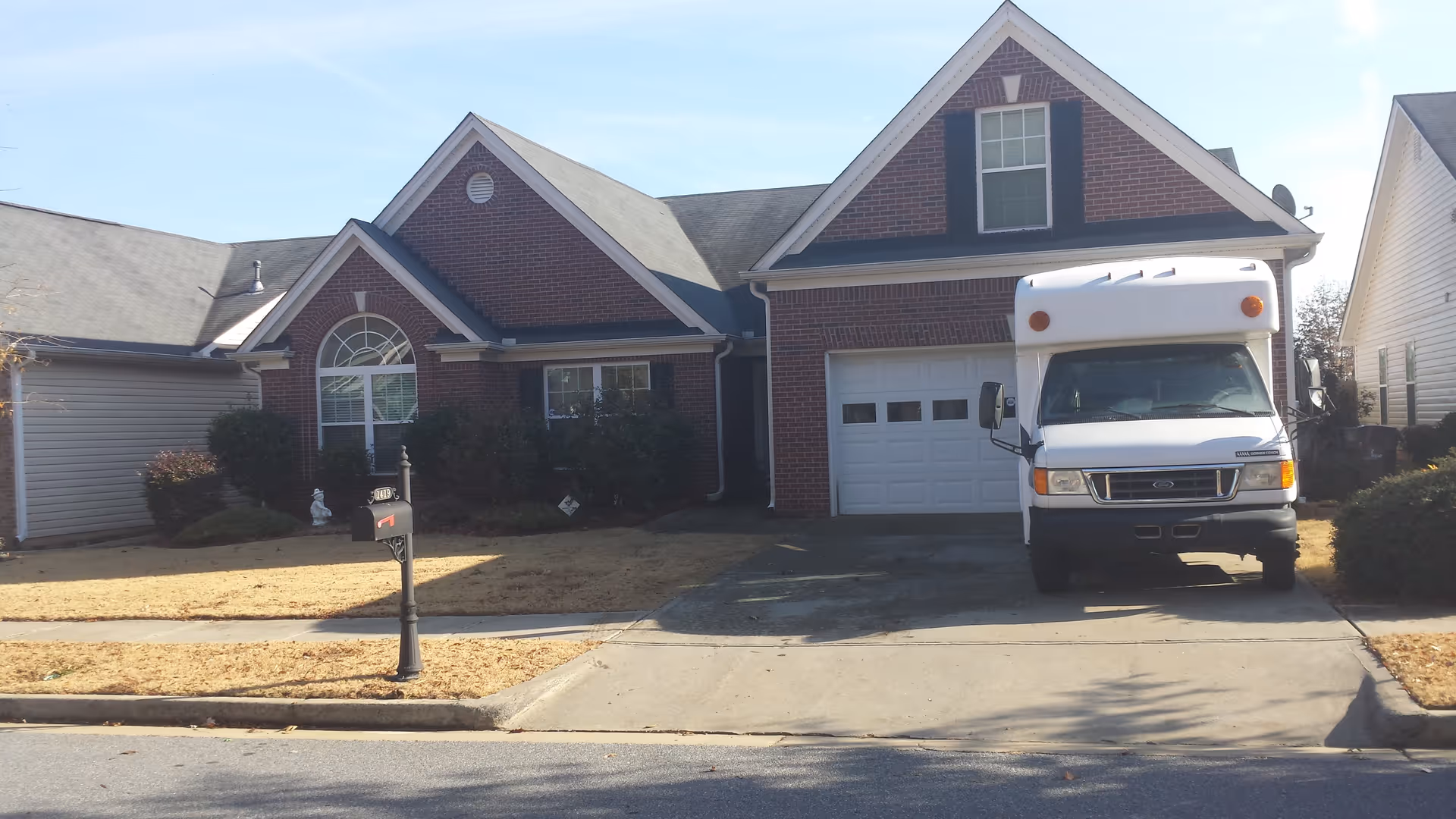 Front exterior view of a single-story brick house with a white garage door and a white delivery truck parked in the driveway. The house has a large arched window and black shutters. There is a mailbox on the curb and some bushes in the front yard.