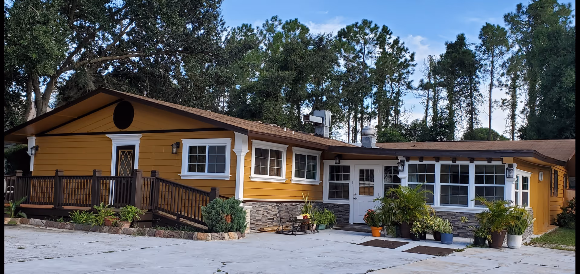 Exterior view of a single-story yellow building with white trim and a brown roof, surrounded by trees. The building has multiple windows, a ramp with railings, and several potted plants near the entrance.