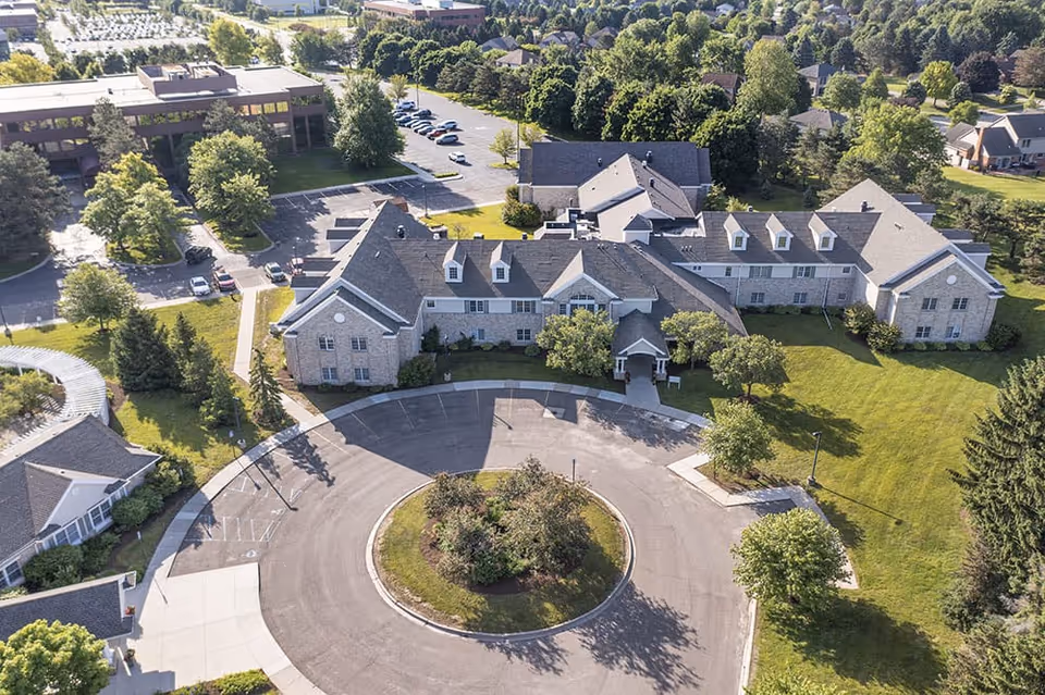 Aerial view of a senior living facility named Brookdale Troy showing a large building with a circular driveway and landscaped greenery surrounding it. There are several trees, parking spaces, and nearby buildings visible in the background.