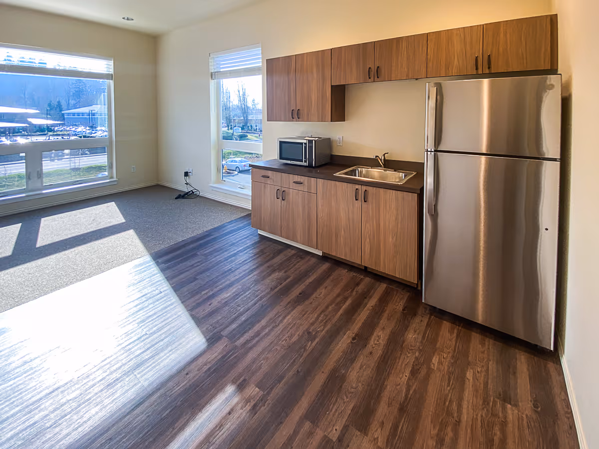 A bright room with large windows letting in natural light, featuring a kitchenette with wooden cabinets, a stainless steel refrigerator, a microwave, and a sink. The floor is a combination of dark wood and carpet.
