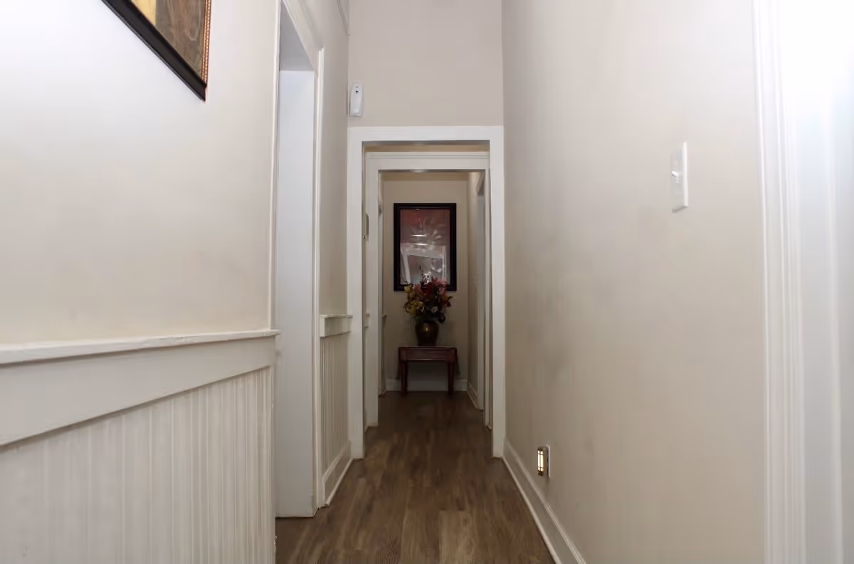 A narrow hallway with light beige walls and wood flooring. There is a small table with a vase of flowers and a framed picture on the wall at the end of the hallway. The hallway has white trim and a light switch on the right wall.