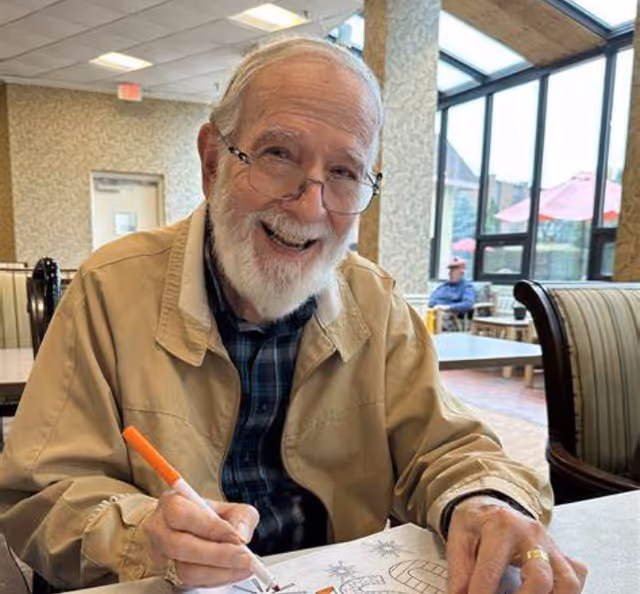 An elderly man with glasses and a white beard is sitting at a table indoors, smiling while coloring a picture with an orange marker. The room has large windows showing outdoor umbrellas and another person sitting outside.