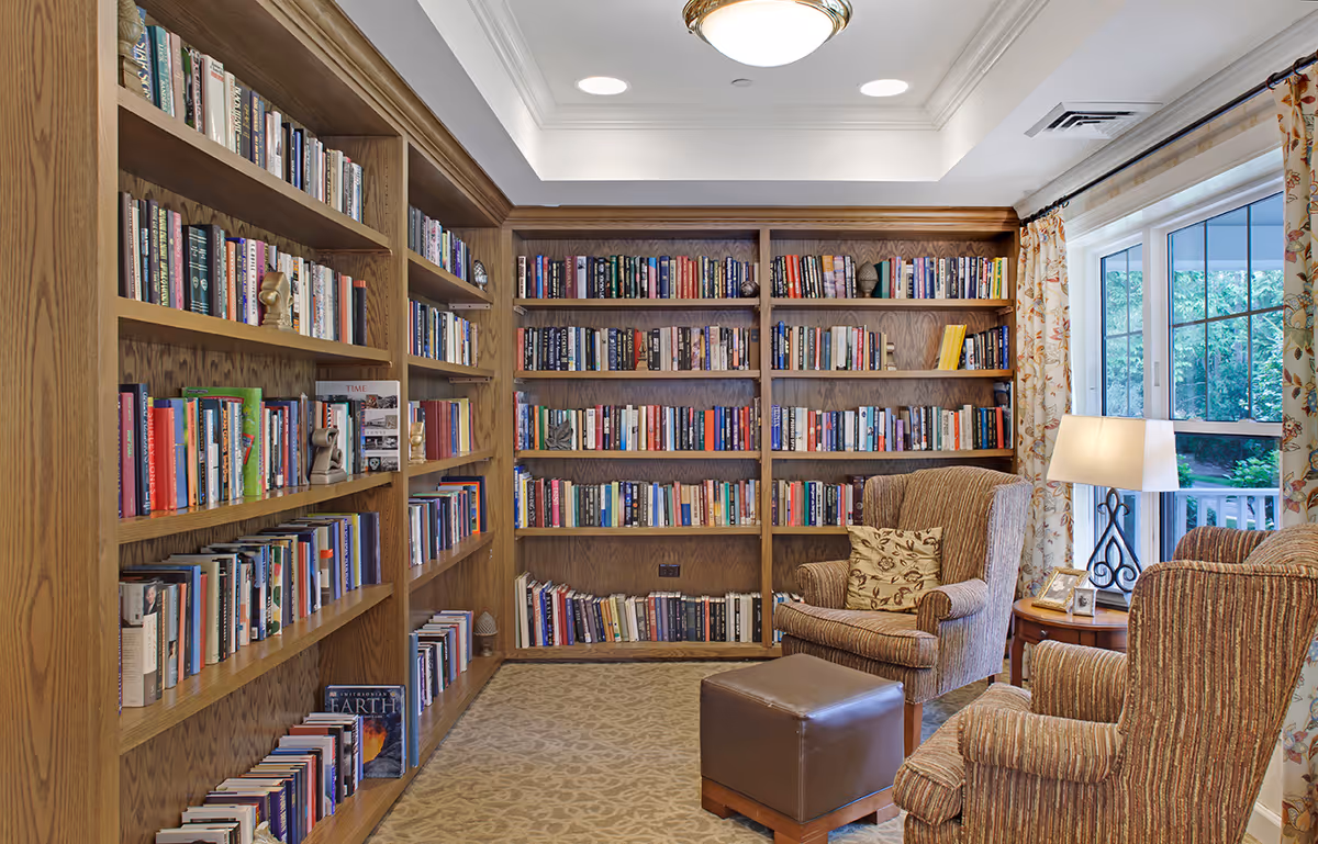 Cozy library with wooden built-in bookshelves, two upholstered armchairs, a lamp, and a window overlooking greenery.