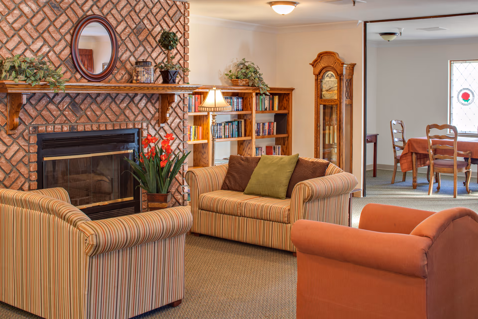 Cozy living room with striped sofas and armchairs arranged around a brick fireplace, a bookshelf and grandfather clock nearby and a dining area visible in the background.