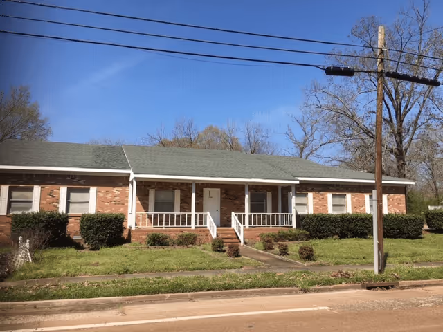 Single-story brick building with a green roof, white front door, and white railings on the front porch. The building is surrounded by green grass, bushes, and trees under a clear blue sky.