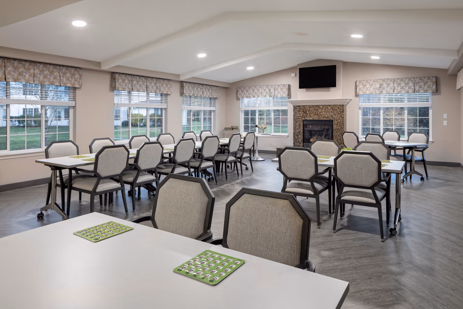 A bright and spacious dining room with multiple tables and cushioned chairs arranged neatly. The room features large windows with patterned valances, a fireplace with a mounted TV above it, and a light-colored floor. Each table has a green placemat with a grid of images or text.