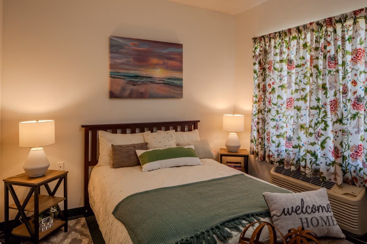 A cozy bedroom with a bed featuring multiple pillows and a green throw blanket. There are two bedside tables with lamps on each side of the bed. A colorful floral curtain covers the window, and a painting of a sunset over the ocean hangs on the wall above the bed. A pillow on a chair in the foreground reads 'welcome HOME'.