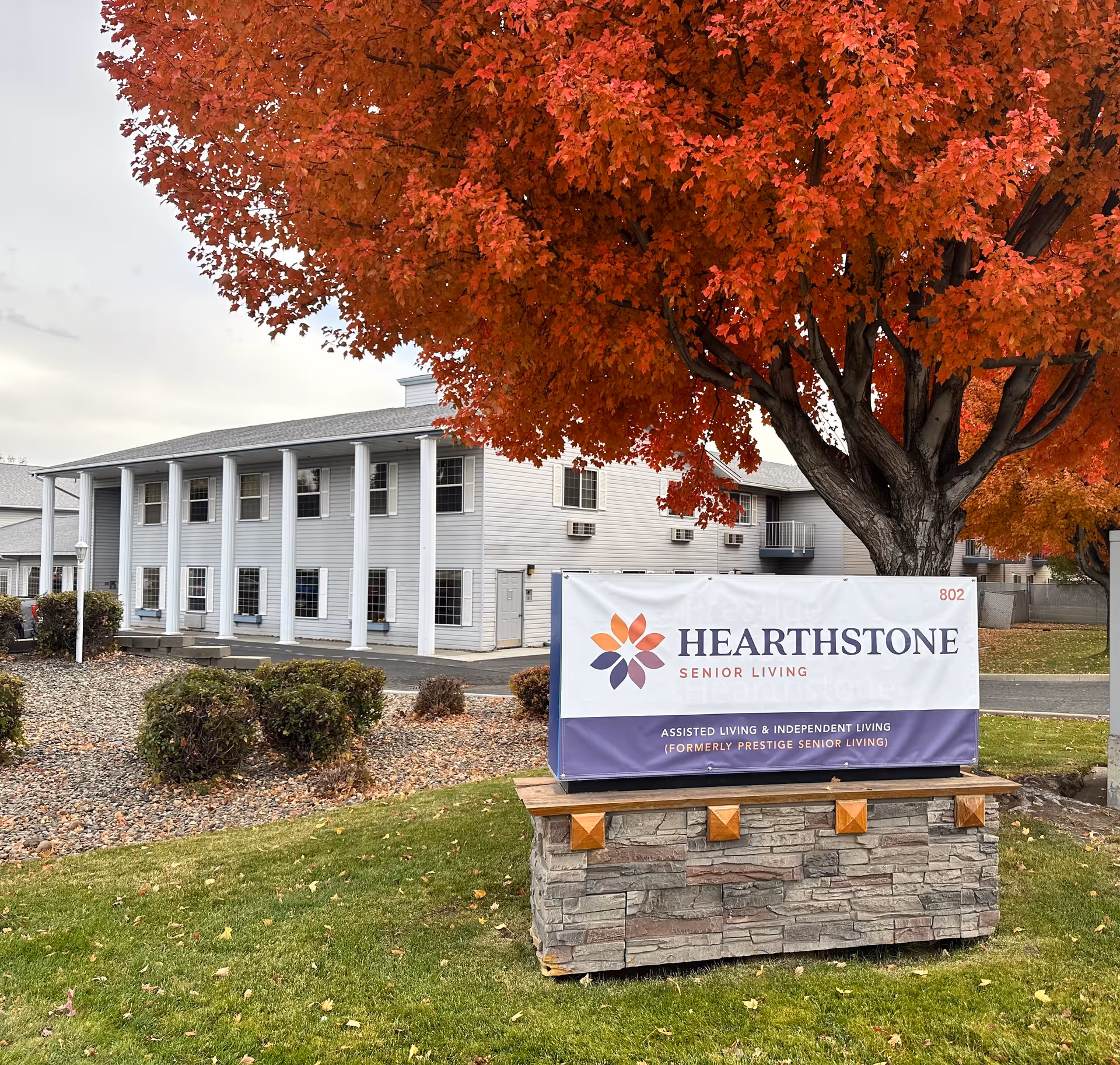 Front of Hearthstone Senior Living building with a large sign and a tree with red autumn leaves in the foreground.