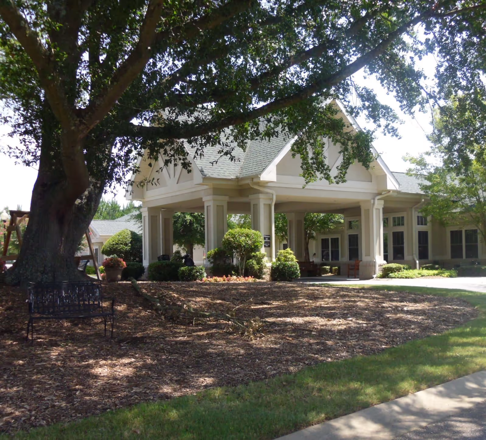 Exterior view of a senior living facility entrance with a covered driveway supported by columns, surrounded by trees, bushes, and a bench under a large tree providing shade.