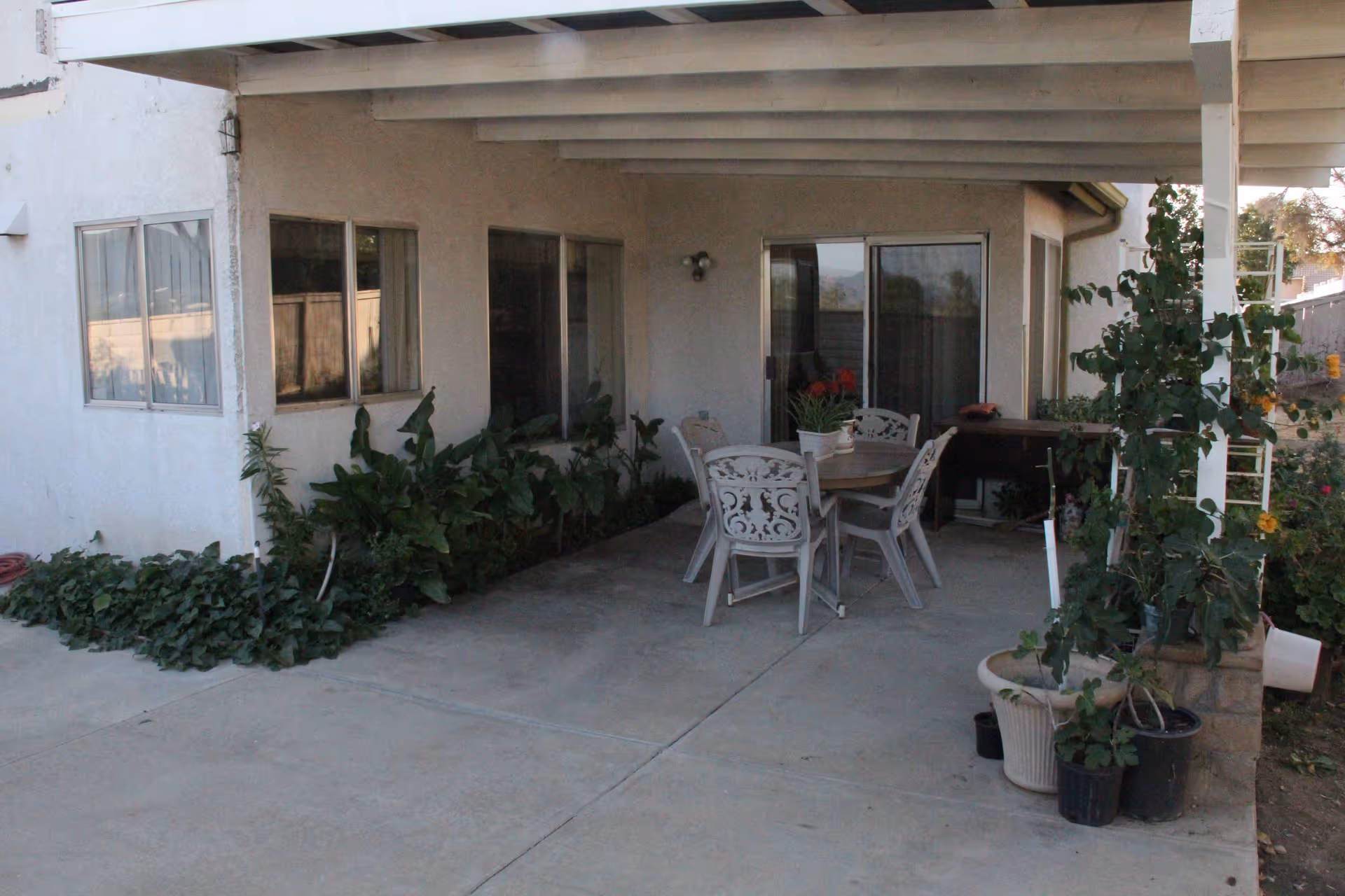 Covered outdoor patio area with a round table and four chairs. There are potted plants and greenery along the edges, with windows and a sliding glass door leading into the building.