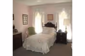 A small bedroom with a single bed covered in a white lace bedspread. The bed has a dark wooden headboard and is positioned against a wall with two windows covered by sheer white curtains. To the right of the bed is a dark wooden nightstand with a lamp and a decorative item. To the left of the bed is a green upholstered chair and a dark wooden dresser with framed pictures on the wall above it. The walls are painted light pink.