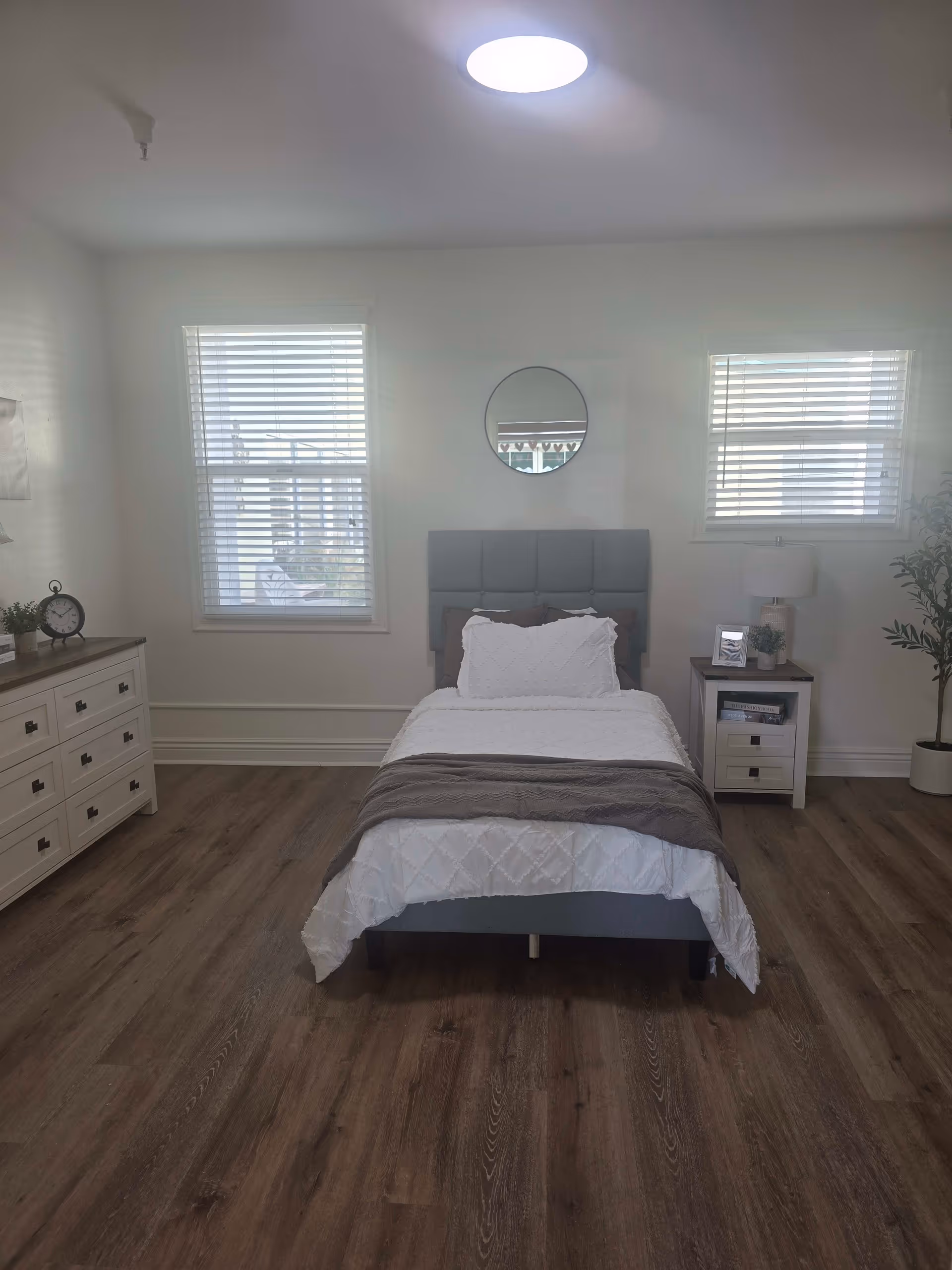 A neatly arranged bedroom with a single bed centered against a wall. The bed has a gray upholstered headboard, white bedding, and a gray throw blanket. On either side of the bed are two windows with white blinds. To the left of the bed is a white dresser with black handles, a small clock, and a potted plant. To the right is a white nightstand with a lamp, a framed photo, a small plant, and some books. A round mirror hangs on the wall above the bed. The floor is wooden with a medium brown finish, and there is a potted plant in the corner on the right side of the room.