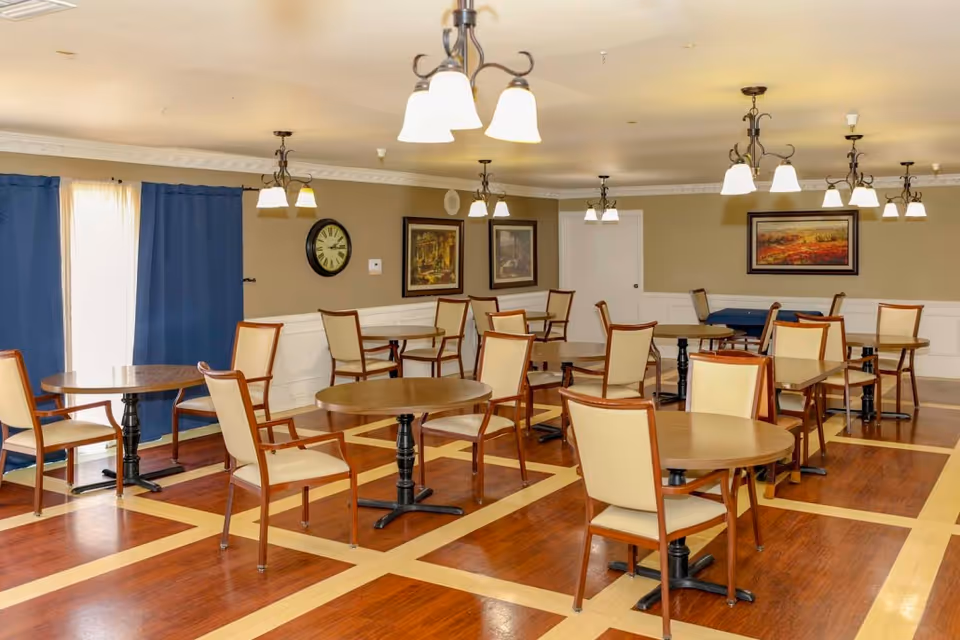 Dining room with multiple round wooden tables and cream-upholstered chairs under hanging chandeliers.