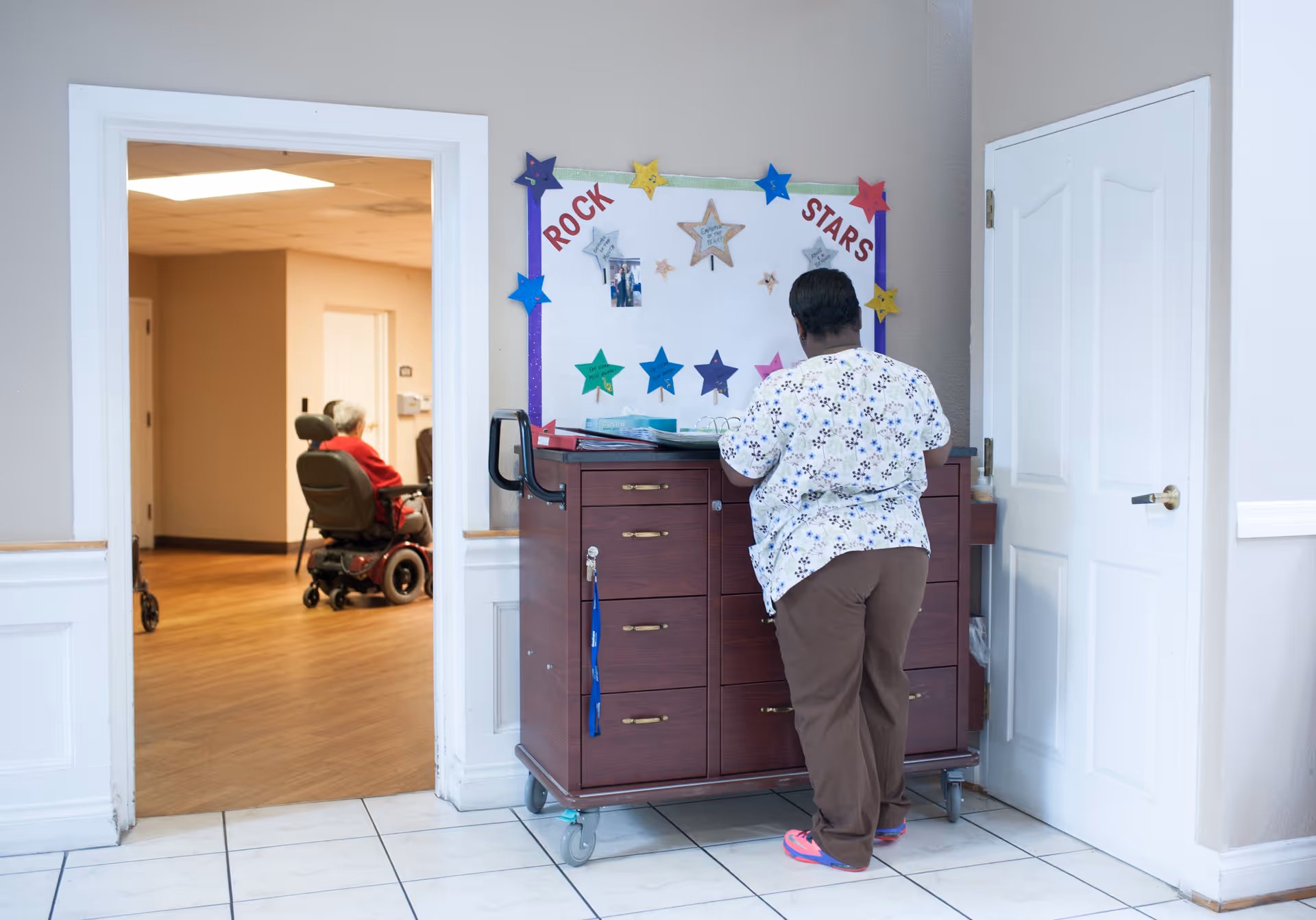 A caregiver in a floral scrub top and brown pants stands facing a wooden cabinet with drawers on wheels, organizing papers in front of a bulletin board decorated with colorful stars and the words 'ROCK STARS'. In the background, an elderly person in a red shirt is seated in a motorized wheelchair in a hallway with wooden flooring.