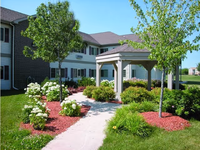 Outdoor garden area at Miller's Senior Living Community featuring a concrete walkway leading to a wooden gazebo surrounded by green bushes, flowering plants, and trees with a two-story building in the background under a clear blue sky.