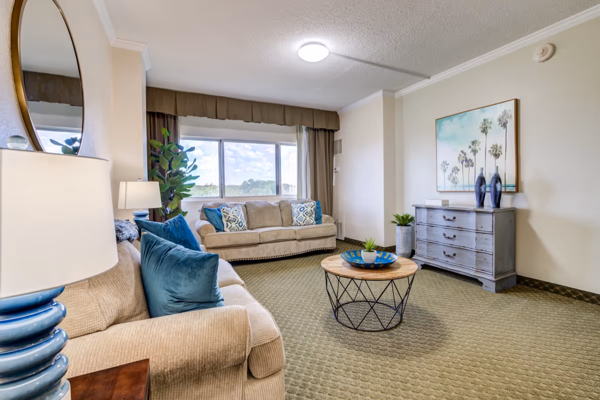 Bright living room with two beige sofas, blue throw pillows, a round coffee table, a dresser with artwork, and a large window letting in natural light.