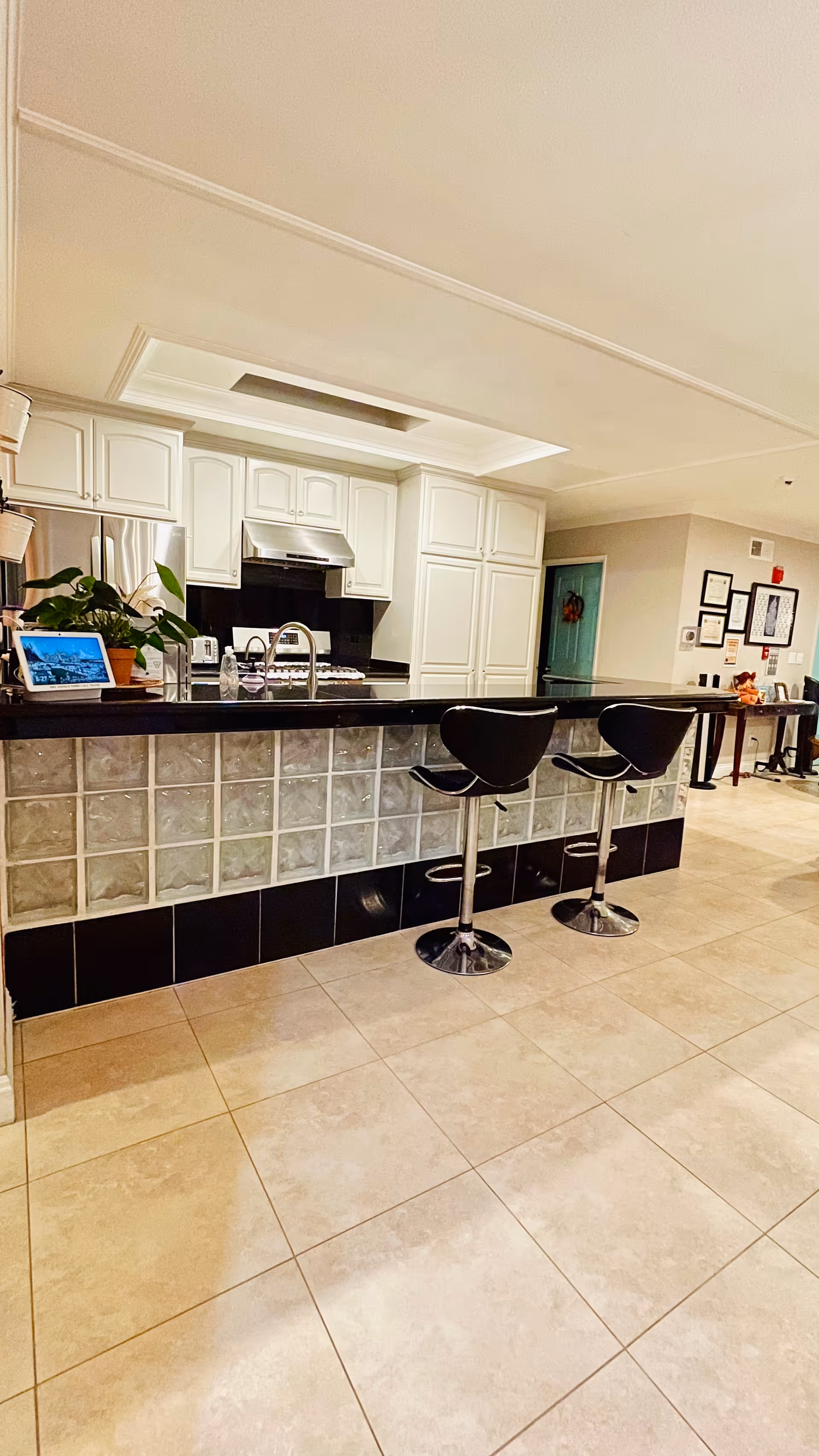 Modern kitchen area with white cabinets, a stainless steel stove and hood, and a black countertop island with glass block base. Two black bar stools are positioned at the island. A plant and a tablet are on the countertop. The floor is tiled and there is a hallway with framed pictures on the wall in the background.