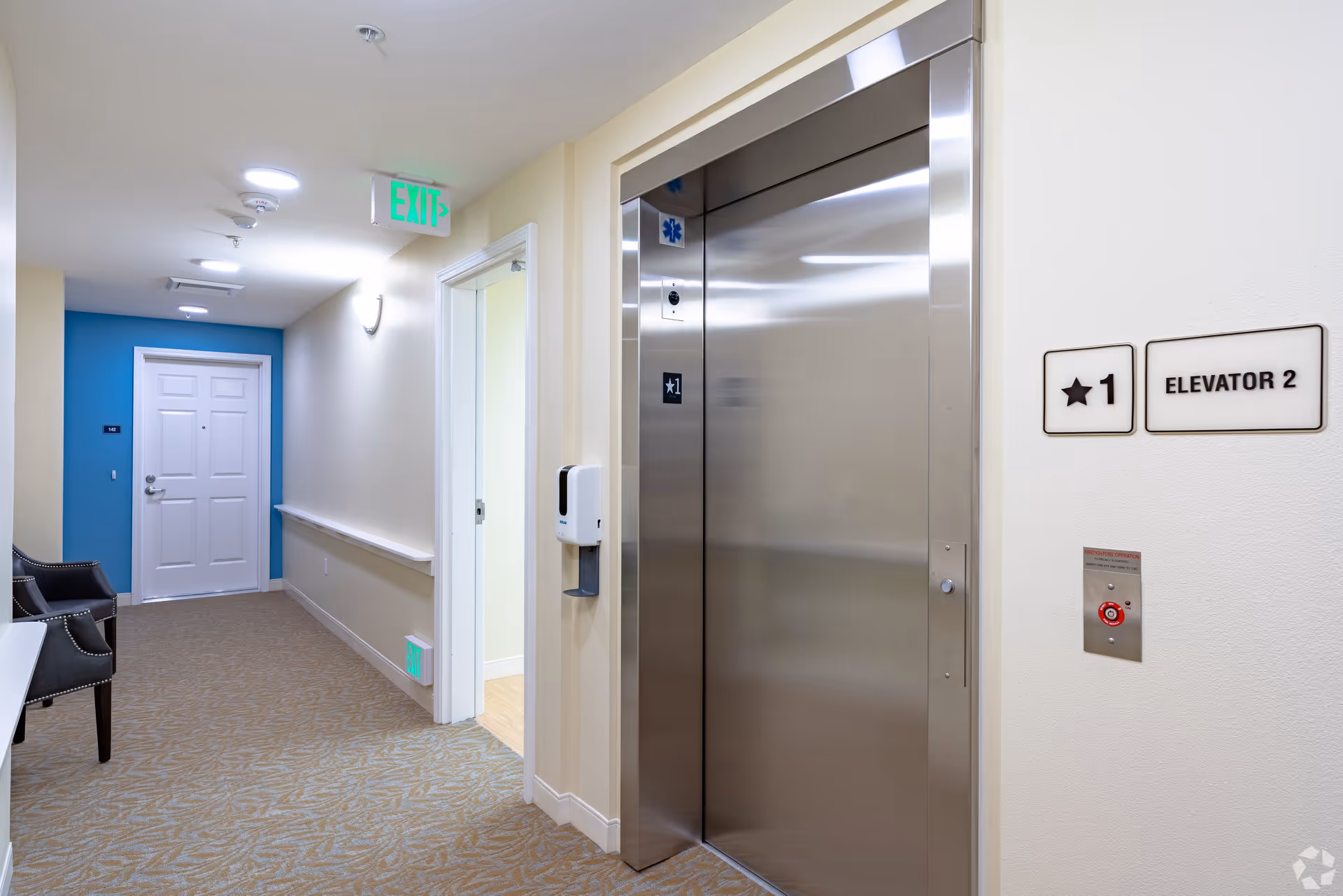 Interior hallway of a senior living facility showing a stainless steel elevator labeled 'Elevator 2', an exit sign, seating, and a doorway.