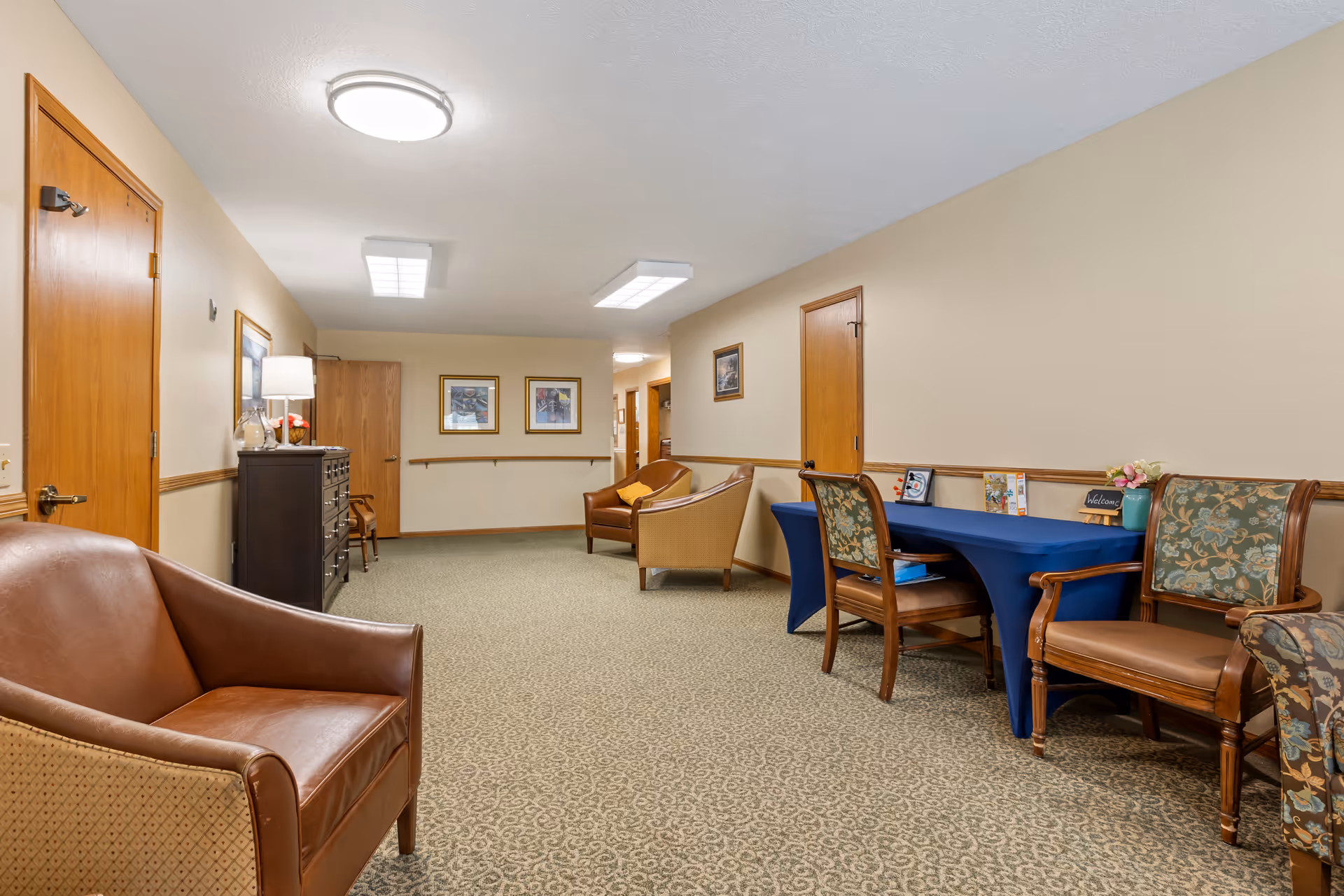 A hallway in a senior living facility with beige walls and patterned carpet. The hallway features several wooden doors, framed artwork on the walls, and a few seating areas with leather and upholstered chairs. A table covered with a blue cloth holds a small welcome sign and decorative items.