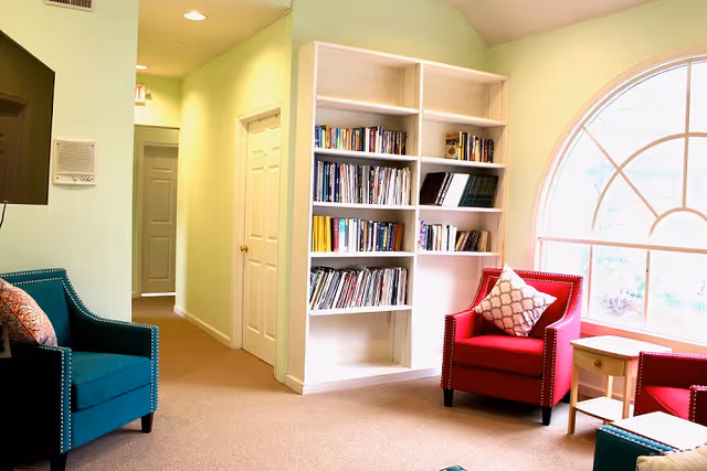 A cozy sitting area in a senior living facility with a large arched window letting in natural light. The room features a white bookshelf filled with books, a red armchair with a patterned cushion, a teal armchair with a cushion, a small wooden side table, and a hallway with doors leading to other rooms.