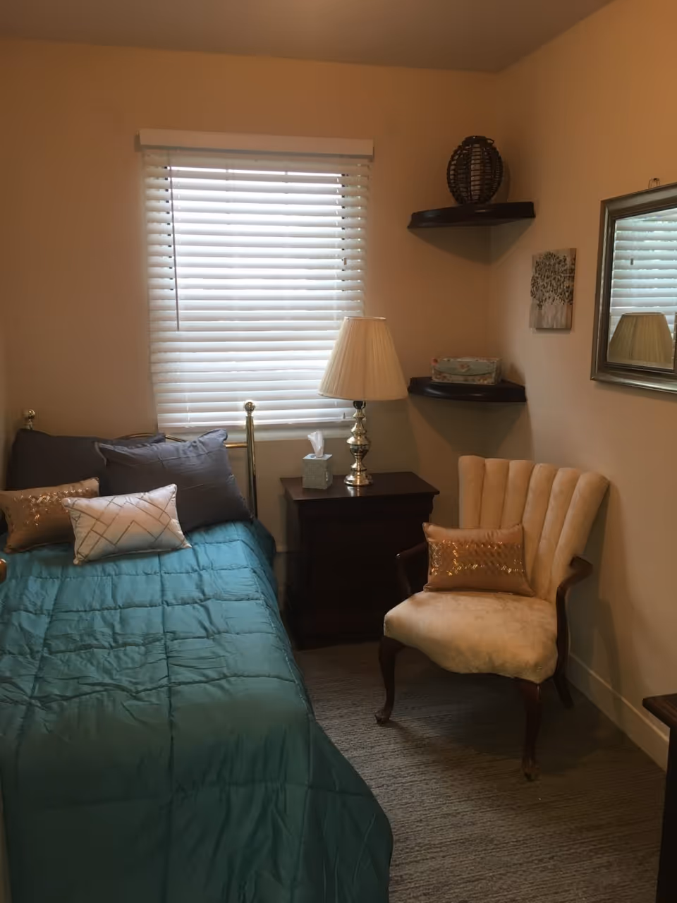 A small bedroom with a single bed covered in a teal comforter and multiple pillows. Next to the bed is a dark wooden nightstand with a silver lamp and a tissue box. A beige upholstered armchair with a decorative pillow is placed near the corner, under two floating corner shelves holding decorative items. A window with white blinds is behind the bed, and a framed mirror hangs on the adjacent wall.
