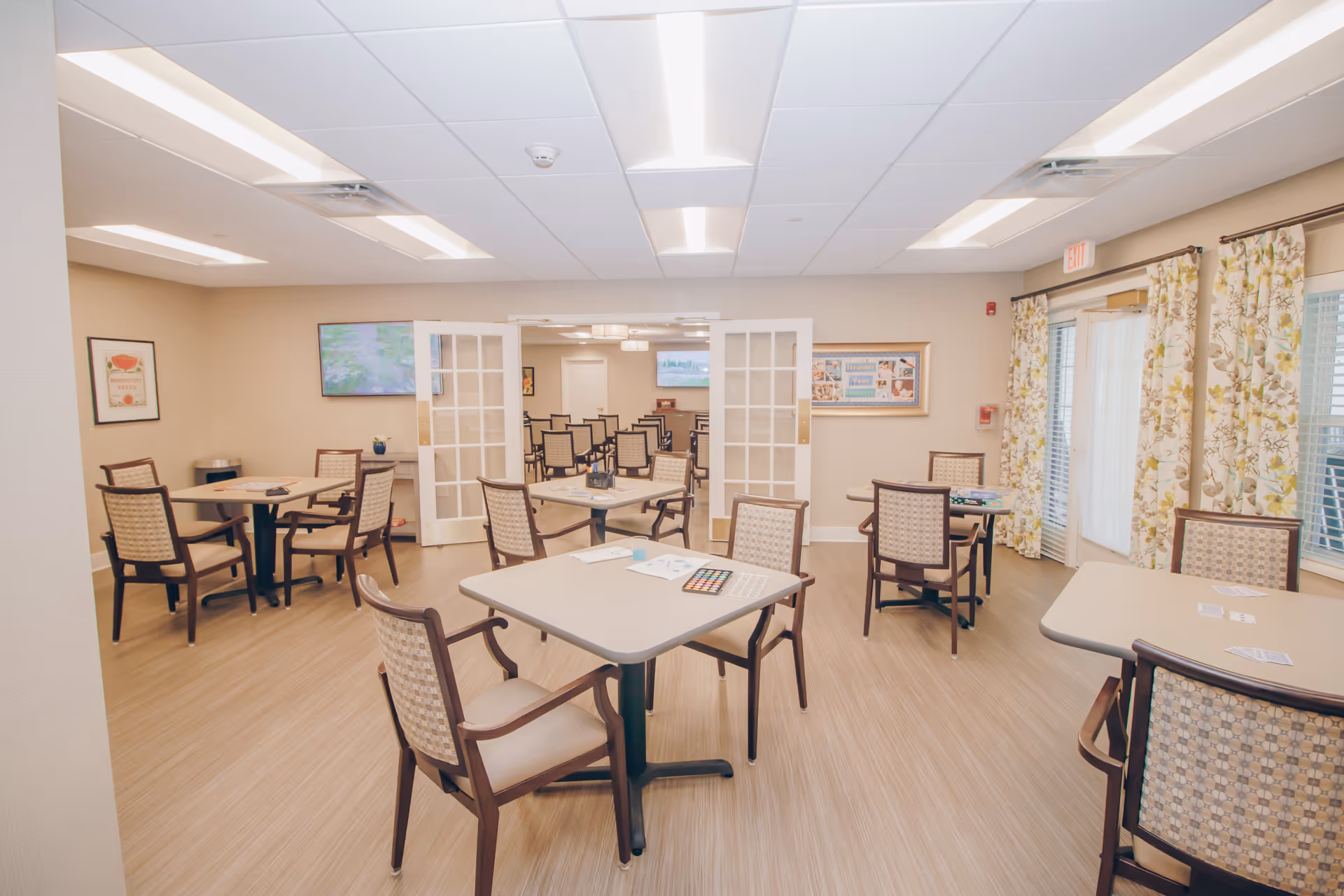 A bright and spacious common area in a senior living facility with several tables and chairs arranged for group activities. The room has light-colored walls, wood flooring, and large windows with floral curtains. There are paintings and a bulletin board on the walls, and a set of double glass doors leading to another room with more chairs and a television.