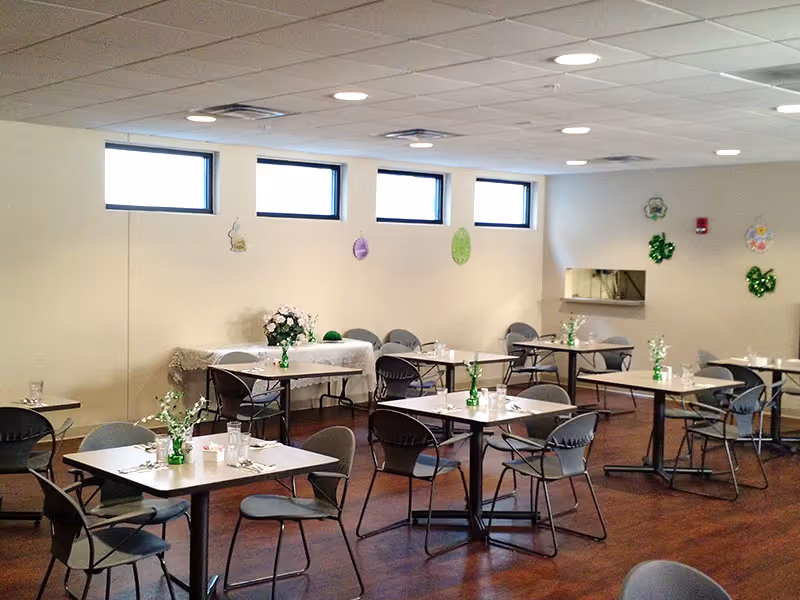 A dining room with several tables and chairs arranged neatly. Each table has a small green vase with flowers and place settings including glasses and napkins. The room has a wooden floor, white walls decorated with hanging ornaments, and small rectangular windows near the ceiling letting in natural light.