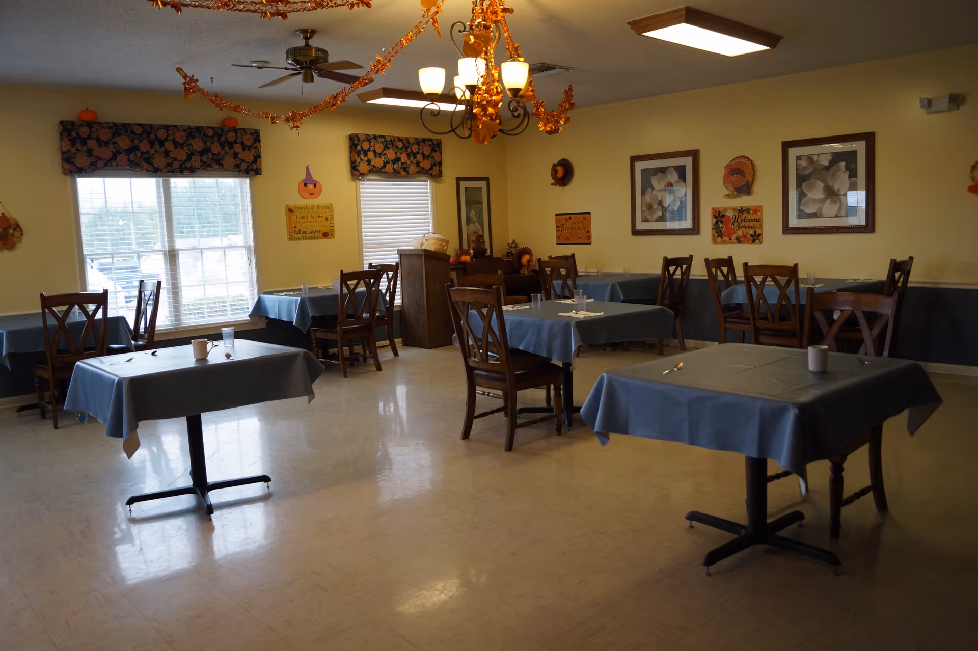 A dining room in a senior living facility decorated with autumn and Thanksgiving-themed decorations. The room has several tables covered with blue tablecloths, each set with cups and utensils. There are wooden chairs around the tables, yellow walls with framed floral pictures, and windows with floral valances. Orange garlands hang from the ceiling light fixtures.
