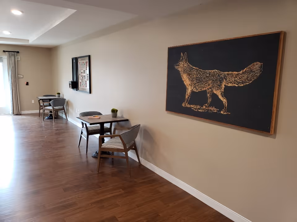 Interior view of a common area with wooden flooring, beige walls, and two small tables each with two chairs. A large framed artwork of a fox is mounted on the right wall. There is a window with curtains at the far end of the room.