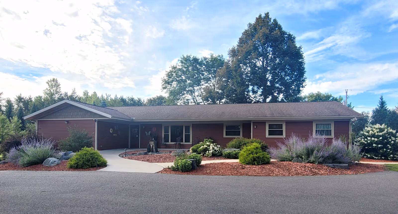 Single-story ranch-style building with a paved driveway, landscaped shrub beds and trees beneath a partly cloudy sky.