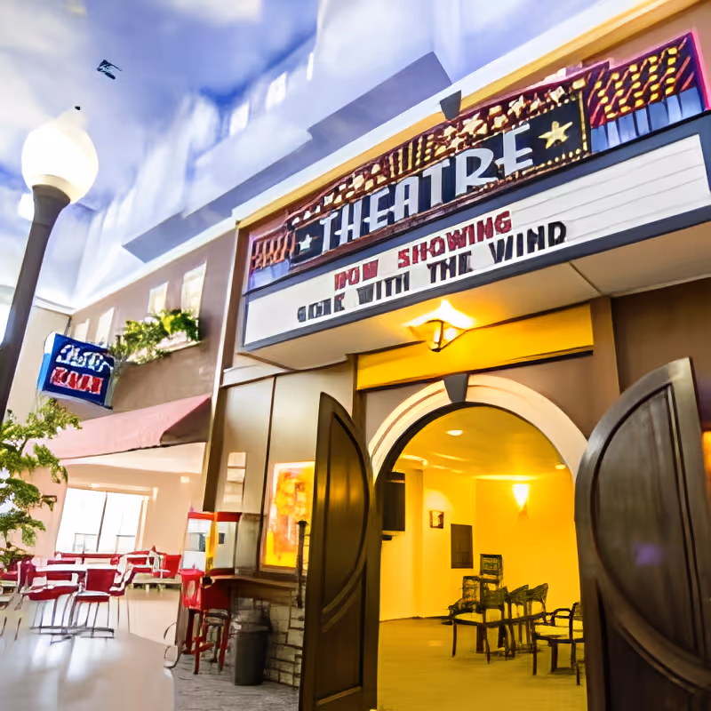Interior view of a small theater entrance with double wooden doors open, a marquee sign above reading 'THEATRE' and 'NOW SHOWING GONE WITH THE WIND'. To the left, there are red chairs and tables in a dining area with a streetlamp and a sign that says 'ASTRO DINER'.