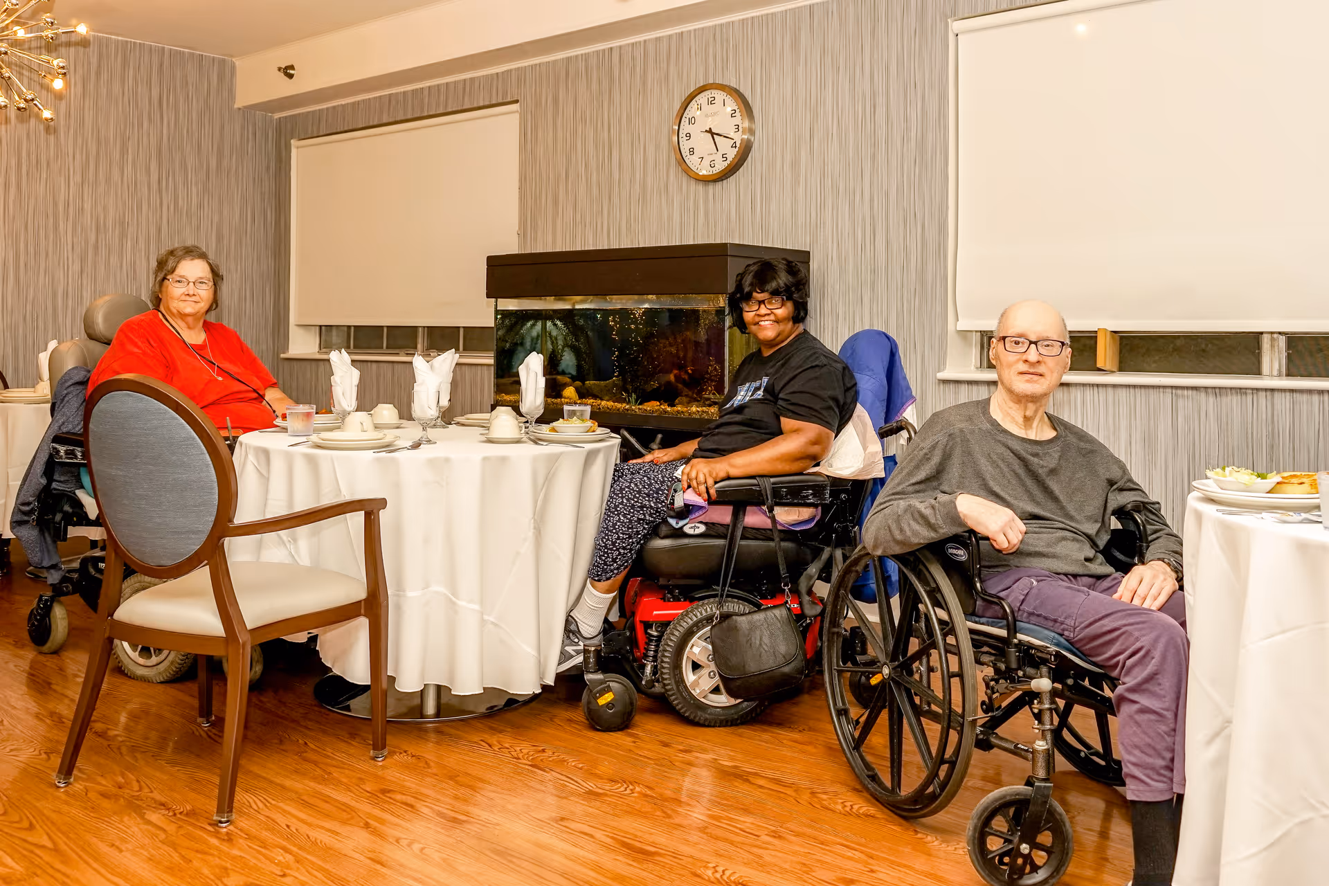 Three elderly residents in wheelchairs seated around a dining table in a dining room with an aquarium and a wall clock.