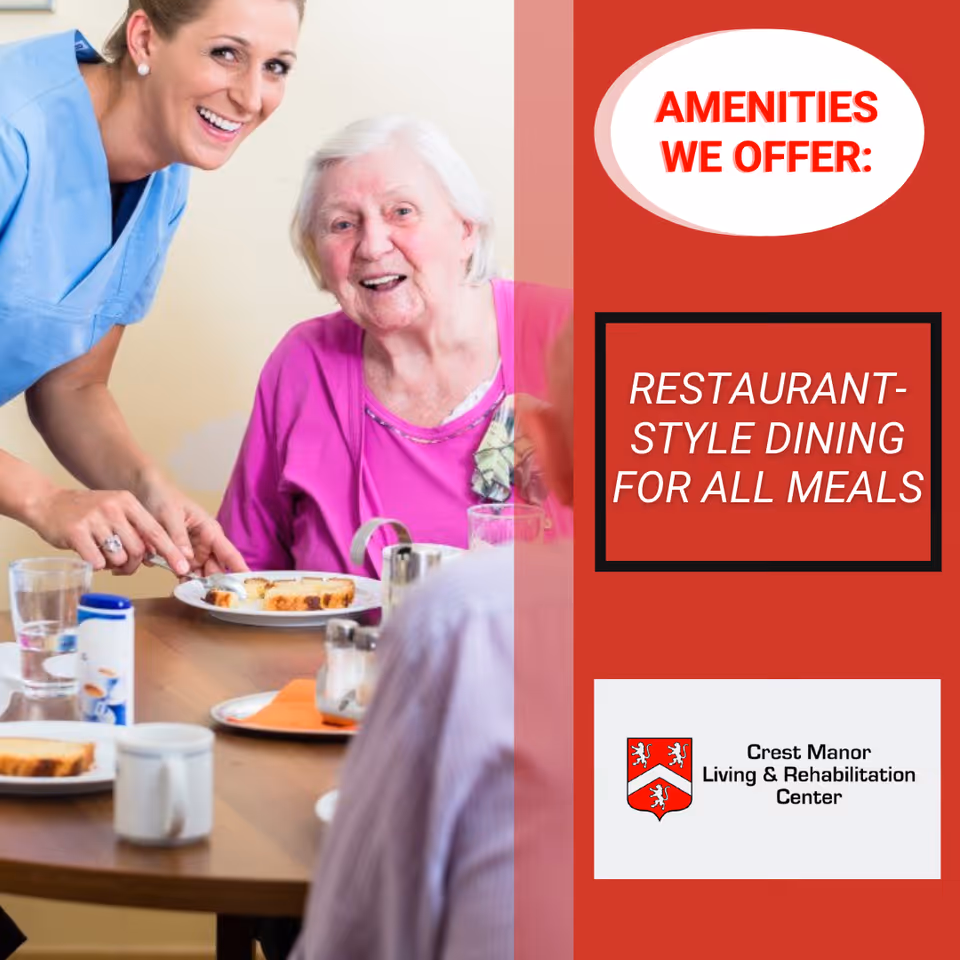 A smiling elderly woman in a pink top sitting at a dining table with food, accompanied by a smiling caregiver in blue scrubs assisting her.