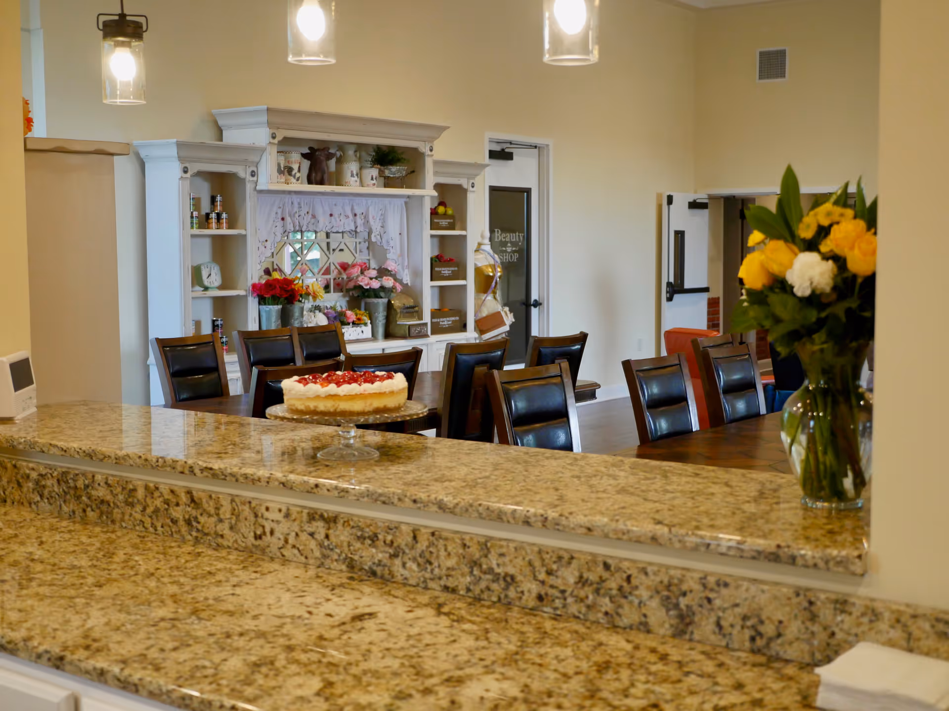 View of a dining area from behind a granite countertop. The dining area has a long wooden table with black cushioned chairs. In the background, there is a white cabinet decorated with various flowers and small items. A door labeled 'Beauty Shop' is visible, and a vase with yellow and white flowers is placed on the countertop.